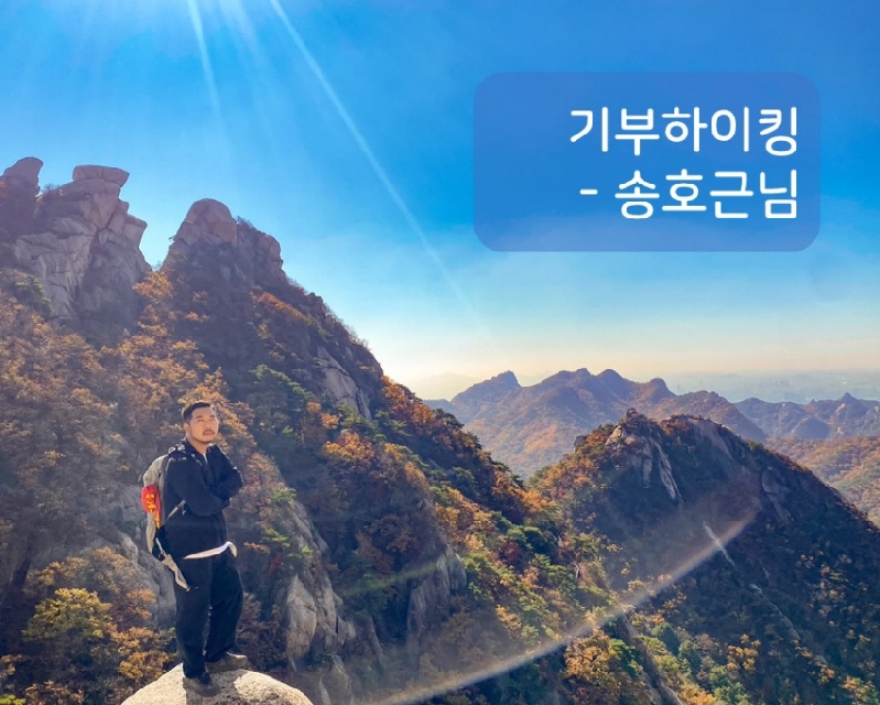 기부하이킹-송호근님, 송호근님이 산 정상에서 서있는 모습, 그 뒤로 파란 하늘에 햇빛이 드리우고 나무와 바위가 어우러진 산의 모습이 보인다.