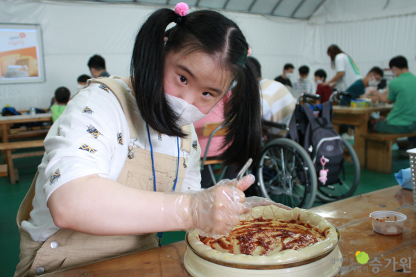 장애가족행복지킴이 승가원ci 삽입 / 피자를 만들고 있는 장애아동 여아. 도우 위 토마토 소스를 바르고 있으며 카메라를 응시하고 있다. 