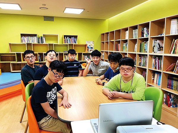 7명의 남성 장애아동이 도서관 책상에 노트북을 중심으로 둘러앉아 영상에 집중하고 있는 모습