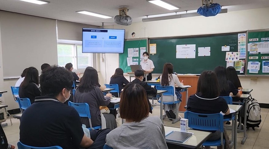 강남대학교 여학생 한명이 고등학교에 가서 사회복지사와 그 업무에 대해 소개하고 있는 모습이다. 앞에는 초록색 칠판이 있고 왼쪽위에 TV스크린이 있다. 학생들은 앉아서 강의를 듣고있다.
