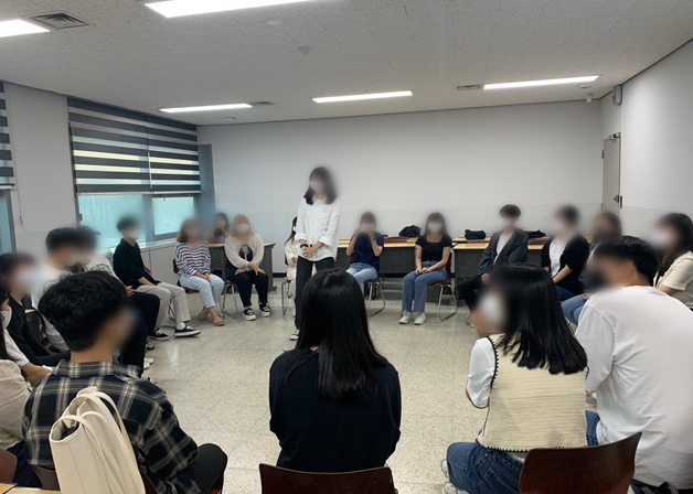 강남대학교 쏘시오 동아리가 극 연습을 하는 모습을 찍은 사진. 약 20명의 학생들이 동그랗게 모여 의자에 앉아 있고 흰색 상의를 입고 검은색 바지를 입은 한 여학생이 원 한 가운데에 서 있다. 다른 학생들은 모두 이 여학생을 바라보고 있다. 
