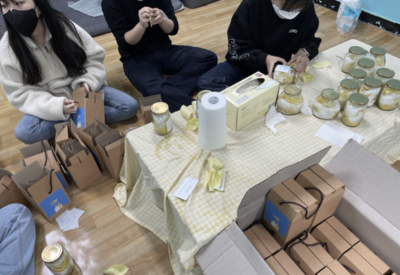 바닥에 앉은 학생 두 명이 레몬청이 담긴 병에 라벨을 붙이고 있고, 다른 한 명은 포장을 하고 있다. 상에는 노란색 식탁보가 깔려 있으며 그 위에 레몬청이 놓여져 있다. 그 뒤에는 포장된 레몬청이 놓여져 있다.