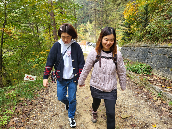 두 명의 여성 이용인들이 산책로를 걷는 모습이다. 두 분 다 미소를 지어 보이며 손을 꼭 잡고 있다.
