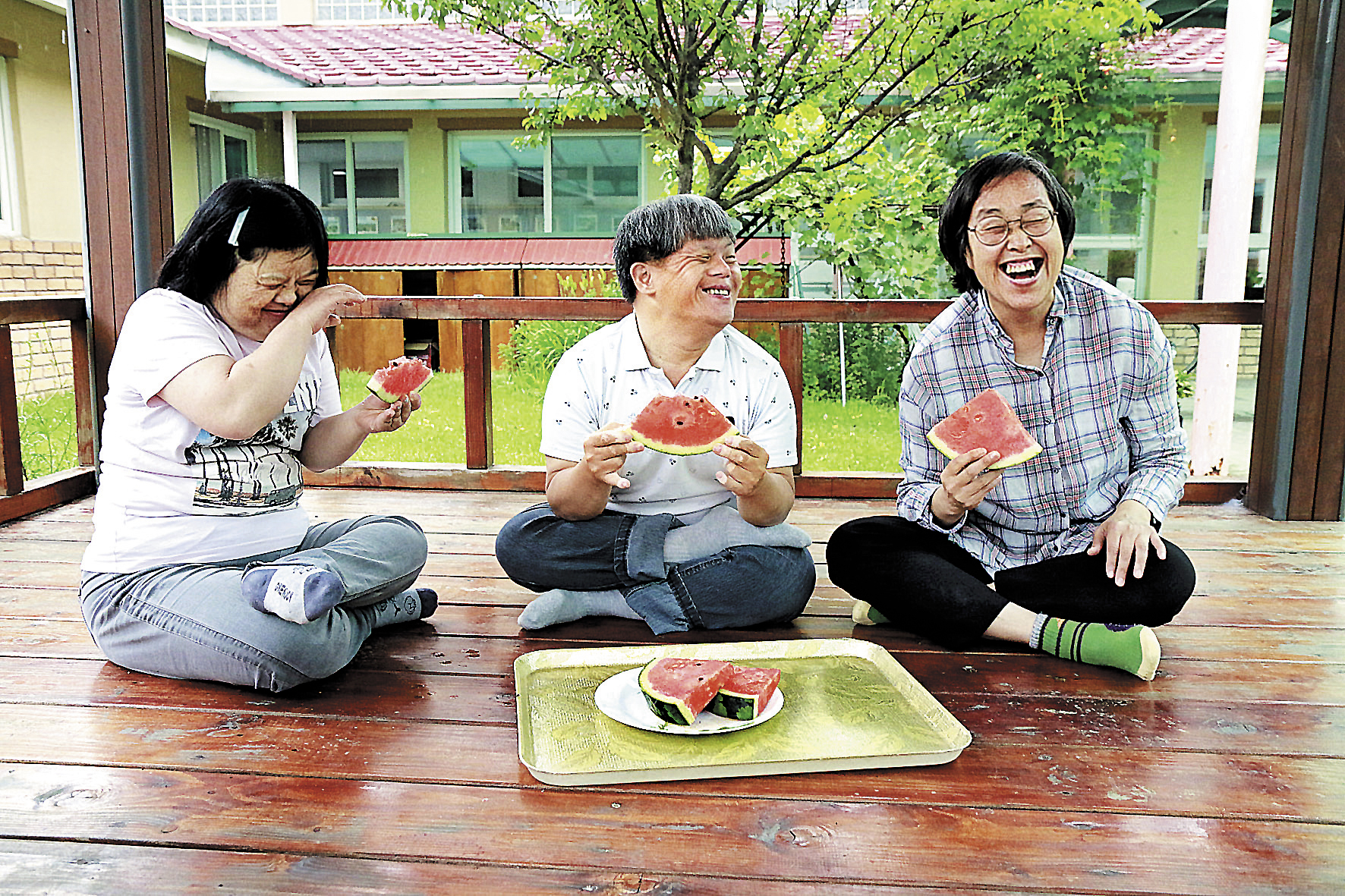 그늘막 나무데크에 앉아서 쟁반에 놓인 수박을 함께 나눠 먹고있는 3명의 장애가족의 모습이다. 얼굴엔 웃음이 가득하다.