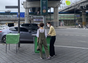 노란색 상의를 입은 여자 사회복지사가 초록색 부스앞에서 멜빵 바지를 입은 시민분께 승가원을 알리는 모습/오른쪽 상단에 장애가족행복지킴이 승가원 CI로고 삽입