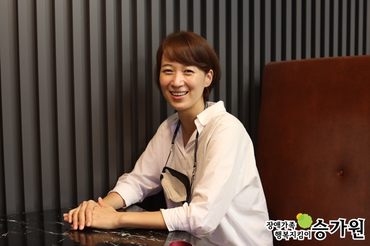 김혜미 후원가족님께서 책상 앞에 앉아 두손을 모으고 환하게 웃으시는 모습, 장애가족행복지킴이 승가원 ci 삽입