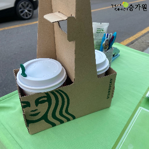 초록색 캠페인 부스 위에는 스타벅스 종이캐리어에 담긴 따뜻한 음료 두 잔이 놓여있음. 오른쪽 상단 위에 장애가족지킴이 승가원 ci삽입