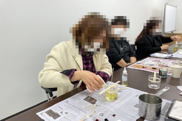 여성 참여자 3명이 테이블 옆에 앉아 캔들만들기를 하고 있다. 테이블에는 신문지가 깔려져 있고 노란빛 투명한 왁스가 있다./ 참가자의 얼굴에는 모자이크가 처리되어 있다.