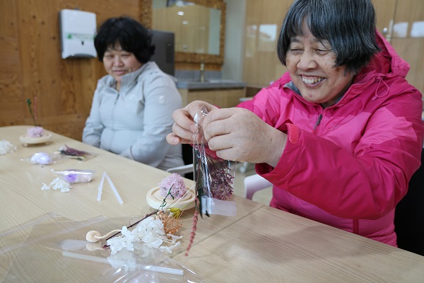 만들기키트 활동에 참여하고 있는 여성이용인 두 명의 모습, 밝게 웃으며드라이플라워를 비닐에서 꺼내고 있다