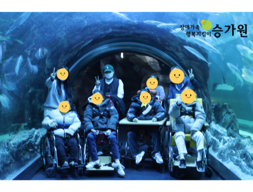 우측 상단에 장애가족 행복지킴이 승가원CI삽입/ 물고기들이 보이는 둥근 유리관 터널 속에 휠체어에 앉은 장애가족 4명과 담당교사 4명이 함께 찍은 사진