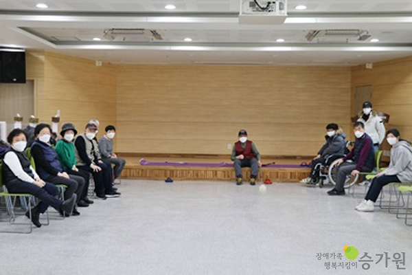 여섯 명의 장애가족이 왼쪽 의자에 앉아있고, 다섯 명의 장애가족이 오른쪽 의자, 휠체어, 가운데 무대에 앉아서 카메라를 응시하고 있는 모습. 모두 마스크를 쓰고 있음. 오른쪽 하단 장애가족행복지킴이승가원CI. 