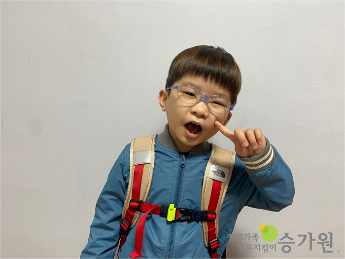 안경을 쓴 장애아동이 왼쪽 손의 검지 손가락을 볼에 콕 찍고 밝게 웃고 있는 모습이다. 오른쪽 하단 장애가족 행복지킴이 승가원 ci 삽입