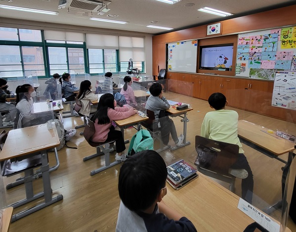 20여명의 초등학생들이 각각 떨어진 책상에 앉아 앞의 화면을 바라보고 있다. 화면에는 캡모자를 쓴 어린이가 나오는 영상이 보인다.
