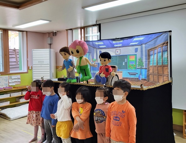 6명의 5세 어린이가 인형극 무대앞에 서서 기념사진을 찍고있다. 인형극 무대에는 연꽃돌이, 가원이, 호동이 인형이 서있다.