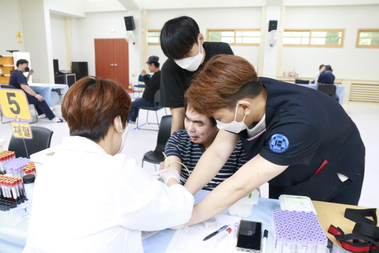 테이블 위에 5/채혈이라고 적혀져 있다. 병원직원이 남자 장애가족의 채혈을 진행하고 있고 두명의 직원이 채혈을 도와주고 있다.