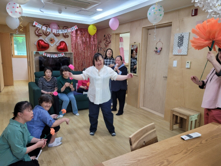 약 9명의 장애가족들이 춤을 추고 노래를 부르고 있다. 한명의 장애가족이 일어서 있고, 나머지 장애가족은 주변에 둘러 앉아있다. 