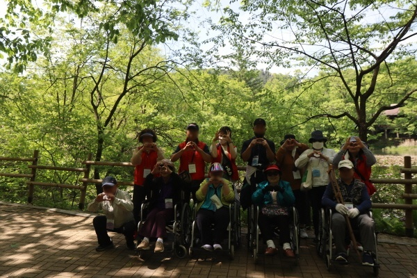 약 12명의 성인장애인나들이 활동의 참여자, 봉사자, 근무가족이 수목원에서 찍은 단체사진이다. 모두가 손으로 하트 포즈를 하고 있다. 뒷편으로 울창한 나무들이 자라있다. 앞의 4명의 장애가족은 휠체어에 타고 있다. 
