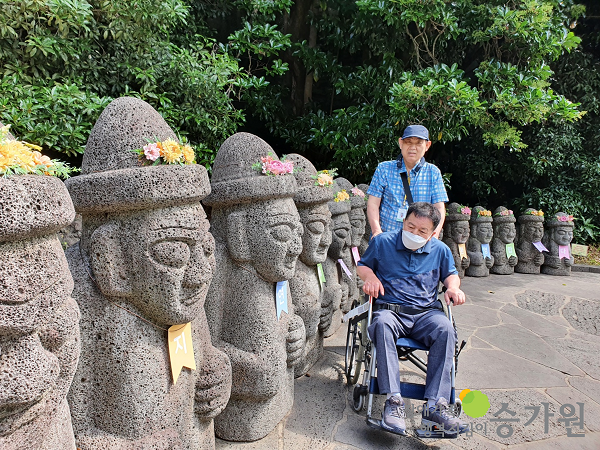 수많은 돌하르방을 배경으로 휠체어를 탄 장애가족과 뒤에서 밀어주는 장애가족의 사진. / 장애가족행복지킴이 승가원ci