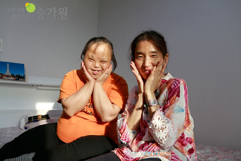 두명의 장애가족이 침대위에서 꽃받침을 한채 환하게 웃고 있는 사진이다./ 장애가족행복지킴이 승가원 CI 삽입