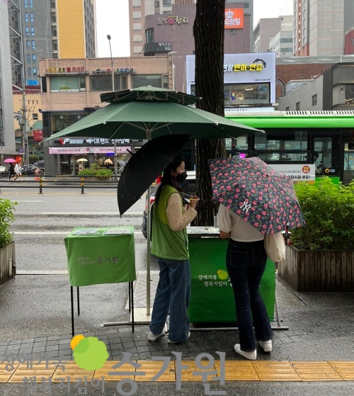 좌측하단 장애가족행복지킴이승가원CI 삽입/비가와서 펼쳐놓은 파라솔 아래에서 여성 사회복지사가 우산을 쓰고 여성 시민분에게 설명 중인 모습. 시민분도 우산을 쓰고 있다.