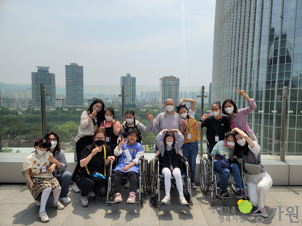 롯데타워 전망대 위에서 장애 아동들과  근무가족, 원장스님이 브이를 하며 밝게 웃으며 단체사진을 찍고 있다./ 오른쪽 하단에 장애가족행복지킴이 승가원CI가 있다.