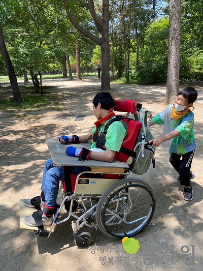 흙길에서 장애아동의 휠체어를 한 장애아동이 열심히 끌어주고 있다. / 오른쪽 하단에 장애가족행복지킴이 승가원CI가 있다