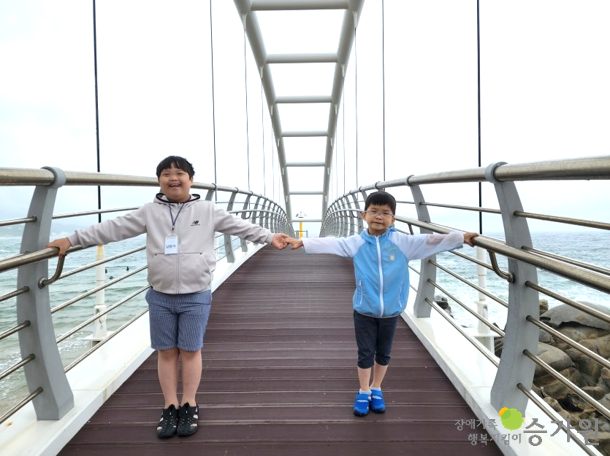 두 명의 장애아동이 다리 위에서 서로 손을 잡고 다른 양팔을 뻗으며 환하게 웃고 있다.
