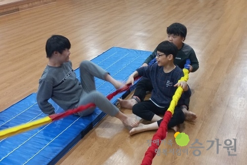 로프를 양쪽 손으로 잡고 있는 남자아이를 중심으로 뒤에 한 명, 왼쪽에 한 명의 남자아이들이 앉아 신체활동을 하고 있다. 오른쪽 하단 승가원ci
