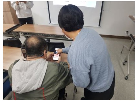 참가자 1명이 의자위에 앉아서 핸드폰으로 어플을 다운로드 하고 있다. 참가자 오른쪽에 교육자 1명이 서서 제로페이 사용 방법을 알려주고 있다.