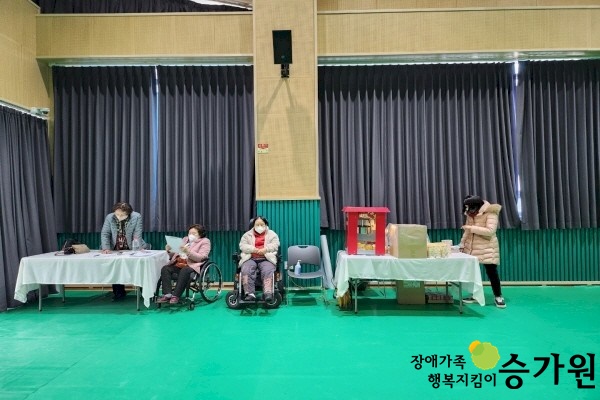 초록색 강당 바닥에 뒤로는 검은색 암막커튼이 쳐져있고 그 앞에 흰 보를 씌운 테이블 위에 박스와 소품이 올려져 있다. 테이블 근처에 2명의 여성은 서있고 두 명의 여성은 휠체어에 앉아 있다. 오른쪽 하단 장애가족 행복지킴이 승가원ci