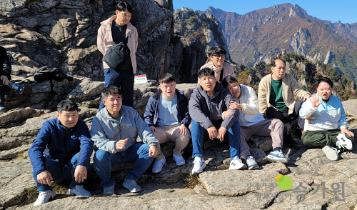 9명의 남성 장애가족들이 산중 돌에 나란히 앉아 사진을 찍은 모습, 우측하단 장애가족행복지킴이 승가원 CI 로고 삽입