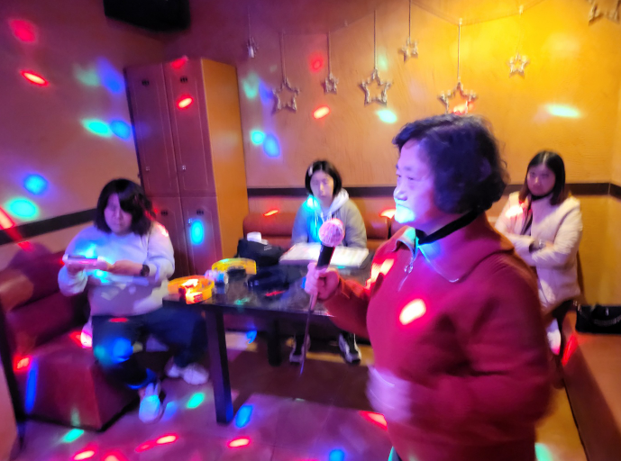 네 명의 여성이 노래방에서 즐겁게 즐기는 모습. 한 명은 일어나서 마이크를 잡고 있고, 한 명은 앉아서 노래를 예약하고 있다.