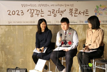 2023 꿈꾸는 그림책 북콘서트라고 적힌 현수막이 걸린 무대위에 작가 세명이 나란히 앉아 있고 가운데 교복을 입은 김수현 작가가 자기소개를 하고있다.오른쪽 아래 승가원 ci 