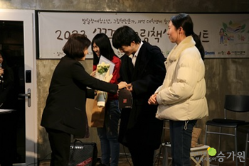 황성혜 관장님이 무대에서 세명의 지역대학생들에게 케이크와 꽃다발을 전달하고 있다.  오른쪽 아래 승가원 ci