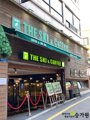 더 스키 카페의 외관. 맨 위에는 초록색 영어 글씨로 'THE SKI & Coffee'라고 적혀져있는 간판이 있고 밑에는 초록색 그늘막이 설치되어 있다. 그 밑에는 영어로 커피 앤 더 스키라고 써 있는 초록색 긴천이 있다. 밑 간판은 형광 연두색 글씨로 'THE SKI & COFFEE'라고 적혀 있고, 글자 양쪽에 더 스키 카페의 로고가 붙어 있다. 넓은 지하로 들어가는 입구에는 판넬이 세워져있고, 빨간색 입구 차단봉이 설치 되어 있다. 가게의 외벽은 모두 나무로 되어 있으며, 카페 입구는 조명이 설치되어 있고, 벽은 통유리창으로 되어 있다. 우측하단 장애가족행복지킴이 승가원 CI