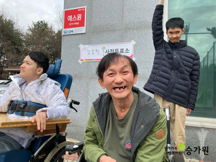장애가족 3명이 투표를 마치고, 사전투표소 앞에서 인증사진을 찍고 있다. 가운데 있는, 연두색 장애가족이 아주 환하게 웃고 있는 모습이다. 사진 하단 오른쪽에 장애가족 행복지킴이 승가원 ci로고 삽입