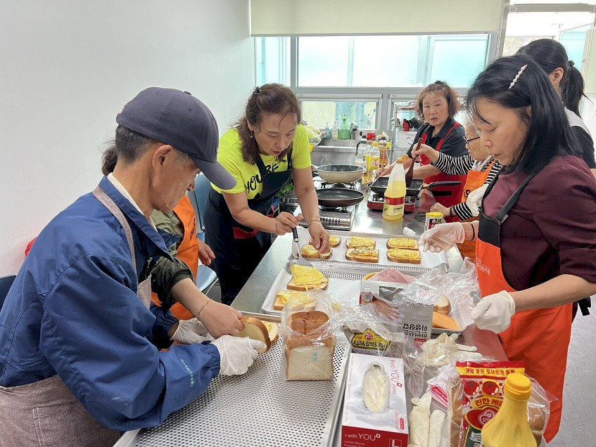 성북구의 지역주민들이 성북장애인복지관에 모여서 요리활동을 진행하고 있다. 샌드위치를 만들기 위해 식빵 위에 계란부침을 얹고 있는 모습. 주민들은 장갑을 끼고 있거나 뒤집개를 들고 있으며 모두가 앞치마를 매고 있다.